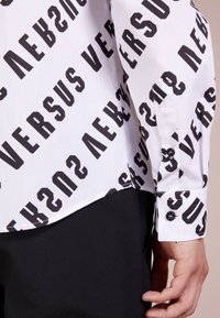 Witte shirt met groot zwart "VERSUS" tekstpatroon. Heeft een knoopmanchet, gladde stof en een ronde zoom. Zwarte stof eronder is zichtbaar.