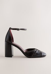 Chaussure à talon haut en cuir noir avec bride à la cheville, bout arrondi et détail découpé sur fond neutre lisse.