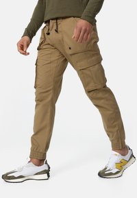 Khaki cargobroek met elastische boorden, meerdere zijzakken en een taille met trekkoord. Draagt witte en groene sneakers met gele accenten.