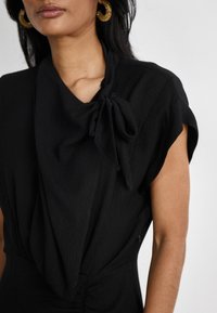 Robe noire avec un devant drapé texturé et un détail en nœud à l'épaule. Avec des manches courtes et des fronces à la taille.