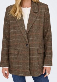 Femme portant un blazer marron à motif chevron avec un seul bouton noir, sur un haut blanc boutonné et un jean bleu.