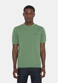 Maglione a maniche corte verde a maglia con colletto e polsini a coste, caratterizzato da un piccolo logo sul lato sinistro del petto. Texture liscia, vestibilità rilassata.