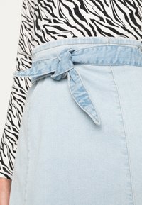 Saia de ganga azul clara com cintura cintada e detalhe de laço, textura suave e costura nas laterais. Top em estampado zebra preto e branco acima.