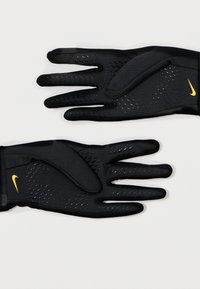 Fekete sportkesztyű, textúrált anyagból, sárga Nike logóval, tapadási mintázattal és passzos csuklódizájnnal.