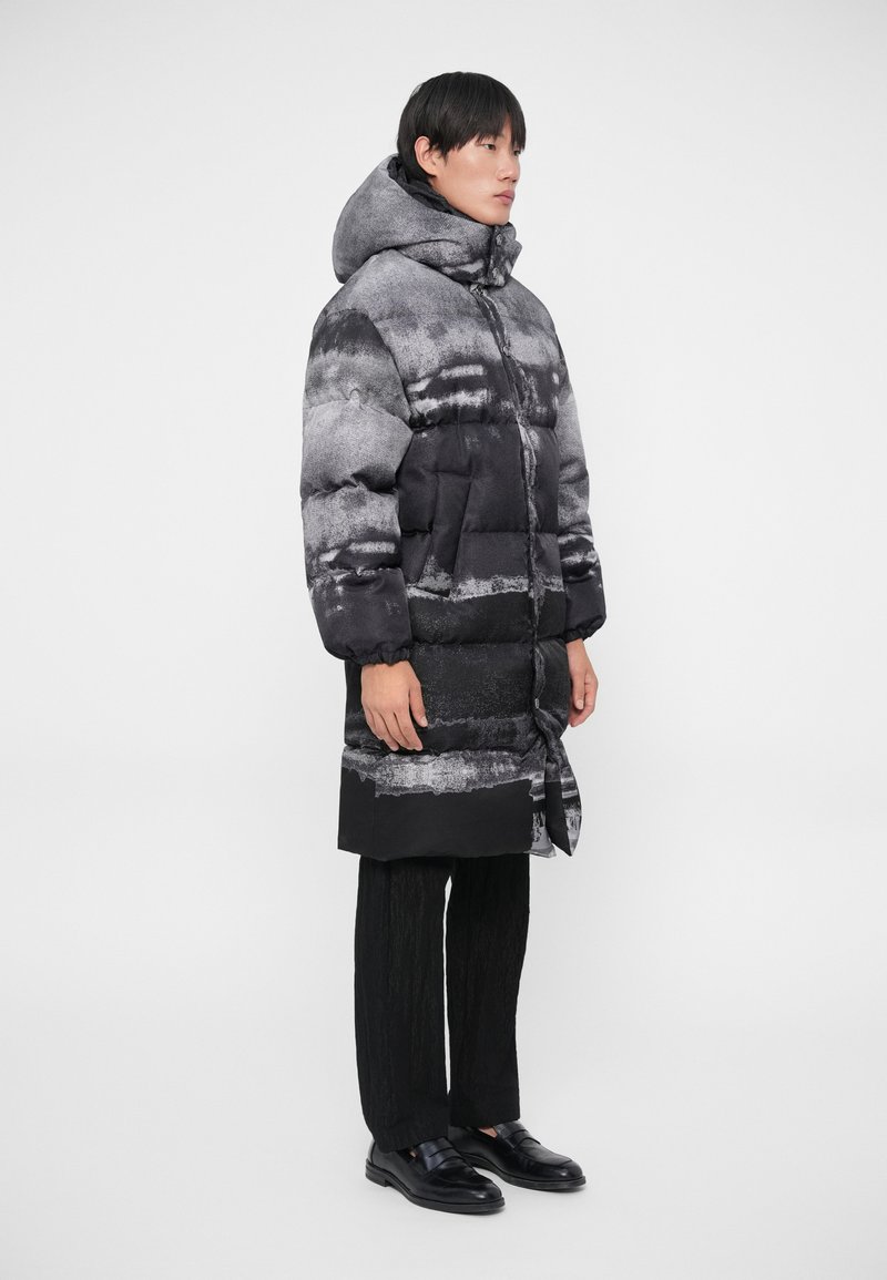 Pufferjakke i sort og grå med et struktureret abstrakt mønster, hætte, frontknapper og sidelommer; parret med sorte strukturerede bukser og sko.