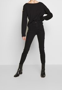 Schwarzes Langarmoberteil mit weitem Ausschnitt und Knopfdetails, kombiniert mit hoch taillierten schwarzen Skinny-Jeans und glänzenden schwarzen Stiefeletten.