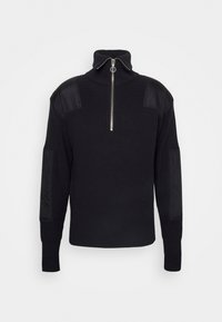 Pull noir en maille avec fermeture à demi-glissière sur le devant, col montant, épaules et coudières matelassées, et manches longues.