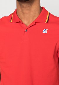 Czerwona koszulka polo z teksturowanej tkaniny, z kołnierzykiem w żółto-granatowe paski oraz logo K-Way na lewej piersi.