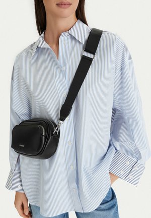 Mujer vistiendo una camisa de botones de rayas azul claro con un bolso cruzado negro con una correa ajustable y bolsillos con cremallera.