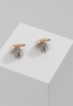 Paire de boucles d'oreilles clous en or rose et argent reposant sur un cube blanc sur fond gris clair.