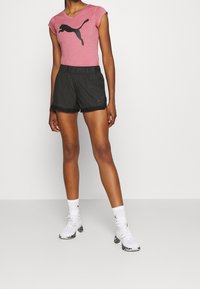 Sportowy zestaw Puma składający się z różowej dopasowanej koszulki z czarnym logo oraz czarnych szortów. Uzupełniony białymi sneakersami z wzorzystą podeszwą.