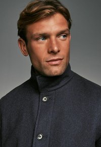 Marineblaue Fischgr�ätjacke mit hohem Kragen, ausgestattet mit zwei schwarzen Knöpfen. Das Material wirkt strukturiert und warm und betont ein modernes Design.