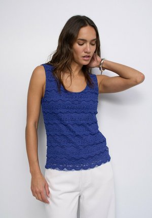 Femme aux longs cheveux foncés portant un haut en dentelle bleue sans manches et un pantalon blanc, posant avec une main derrière la nuque sur un fond blanc.