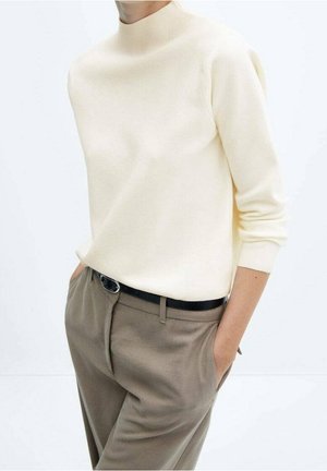 Personne portant un pull à col roulé crème rentré dans un pantalon beige avec une ceinture noire, debout les mains dans les poches.