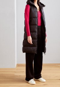 Czarna kamizelka puchowa z kapturem, z pikowanym wzorem i zamkiem błyskawicznym. W połączeniu z dopasowaną czerwoną bluzką z długim rękawem oraz czarnymi, szerokimi spodniami.