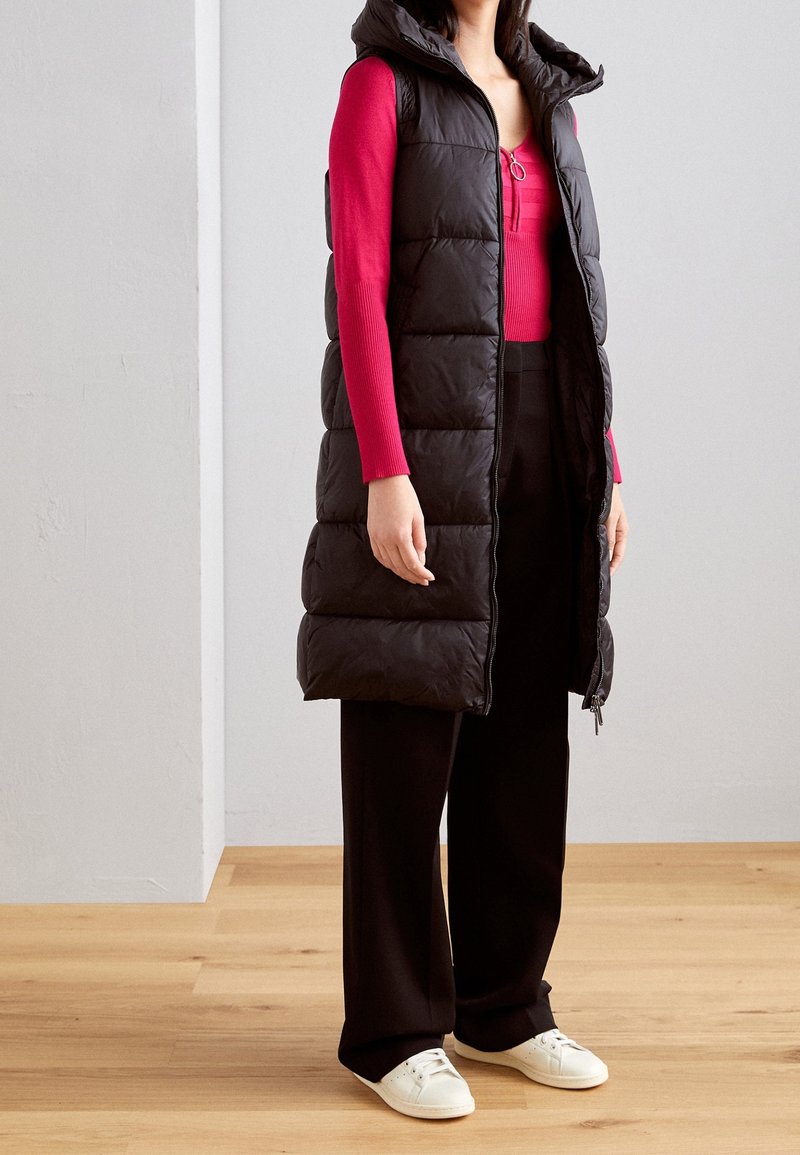 Czarna kamizelka puchowa z kapturem, z pikowanym wzorem i zamkiem błyskawicznym. W połączeniu z dopasowaną czerwoną bluzką z długim rękawem oraz czarnymi, szerokimi spodniami.