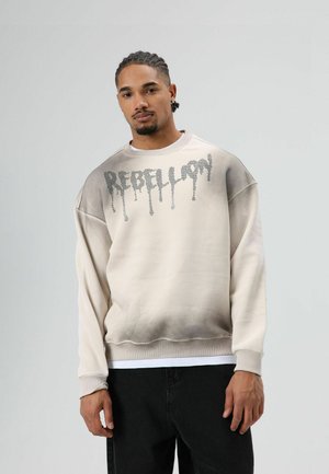 Pullover trui in crème met een gradient grijze onderste helft. Heeft "REBELLION" gedrukt in een druppelend lettertype over de borst.