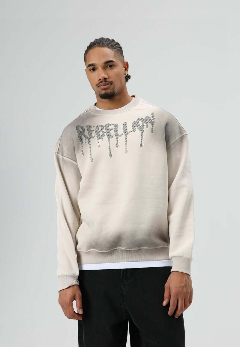 Pullover trui in crème met een gradient grijze onderste helft. Heeft "REBELLION" gedrukt in een druppelend lettertype over de borst.