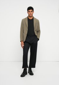 Olijfgroene blazer over een zwarte trui, gecombineerd met zwarte, opgerolde broeken en zwarte combatlaarzen; gladde texturen en een getailleerde pasvorm.