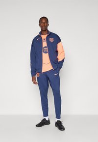 Blå atletisk træningsdragt med koralfarvede detaljer, bestående af en lynlåslukket jakke og tætsiddende bukser, fremstillet af letvægtsmateriale. Sportslogoer synlige.
