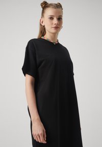 Crna oversize majica od pamučne tkanine. Kratki rukavi, okrugli izrez, ukrašena šarenom ogrlicom od perli za dodatnu pažnju.