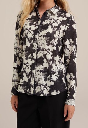 Kvinde iført en sort-hvid blomsterknap-bluse med lange ærmer og sorte bukser, stående mod en ensfarvet baggrund.