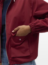 Hånd i lommen på rød jakke med trykknapflap, som viser lynlås, elastisk mansjett, blå genser og denimjeans.