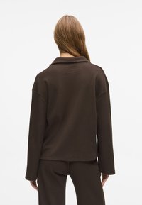 Cămașă lungă cu mâneci lungi, de culoare maro, cu un guler lat și plat și o croială lejeră. Materialul are un aspect texturat și acoperă șoldurile.