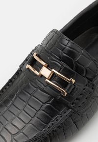 Chaussure en cuir noir au texture crocodile avec une boucle en métal doré sur le devant, comportant des détails cousus autour du bord.