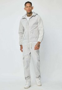 Veste zippée gris clair avec une capuche, dotée de poignets élastiques et de poches latérales ; portée avec un pantalon cargo assorti et des baskets blanches.