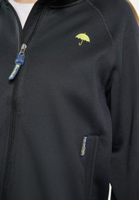 Svart zip-up jacka i texturerat tyg, med ett litet gult paraplyemblem på bröstet och en blå dragkedja.