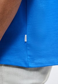 Κοντινή λήψη του τελειώματος ενός μπλε πουκάμισου με μια μικρή λευκή υφασμάτινη ετικέτα και ορατή ραφή, φορεμένο πάνω από ανοιχτόγκρι παντελόνια.