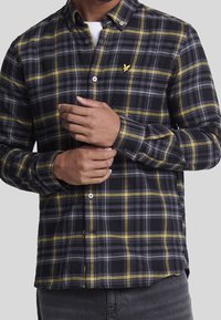 Camisa de franela a cuadros en negro y amarillo con cuello abotonado. Presenta un pequeño logo en el pecho y un ajuste holgado. Tela texturizada.