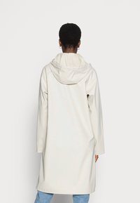 Lång, huvjacka i mjukt, ljusbeige material med en slät yta. Har en avslappnad passform och neddragna axlar, som faller under höfterna.