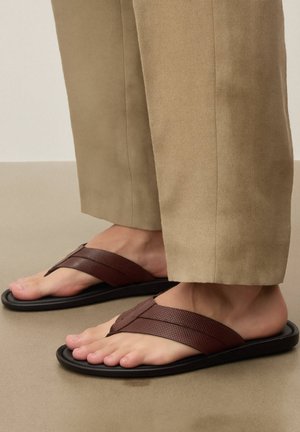 Barna bőr pántos szandált viselő lábak, bézs nadrággal, egy sima, világosbarna padlón állva.