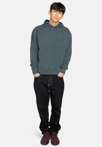 Hooded sweatshirt in muted teal, made of soft fabric, featuring a front pouch pocket. Paired with black jeans and maroon shoes.