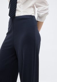 Λευκό πουκάμισο με τα μανίκια αναδιπλωμένα και ριγέ ναυτικό γραβάτα, φορεμένο με ψηλόμεσο, φαρδύ ναυτικό παντελόνι σε ένα άτομο.