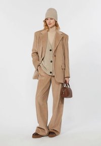 Kvinna i beige kordflorsdräkt, stickad väst, polotröja och mössa, som håller en liten brun läderhandväska, stående mot vit bakgrund.