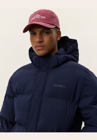 Navy Puffer-Jacke mit hohem Kragen, Kapuze und dem gestickten Logo "LES DEUX" auf der Brust; getragen mit einer burgunderfarbenen Baseballmütze.