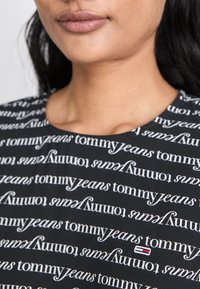 Črn top z ponavljajočim se belim vzorcem "tommy jeans". Opazne podrobnosti vključujejo majhen logo z rdečimi, belimi in modrimi elementi.
