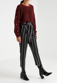 Bordový pletený svetr s perlovými detaily, doplněný černobílými pruhovanými zúženými kalhotami a lesklými černými botami na podpatku.