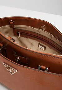 Sac à main en cuir brun texturé avec un compartiment zippé, une plaque logo et un tissu intérieur à motif présentant un design de logo répétitif.