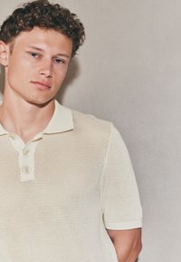 Polo de couleur crème claire, en tissu maille tricoté, avec un col classique et une patte de boutonnage à trois boutons, manches courtes avec poignets côtelés.