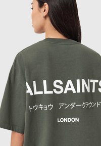 Oliivinvihreä ylisuurikokoinen t-paita, puuvillamateriaalia, jossa on suuri valkoinen teksti takana: "ALLSAINTS", japanilaiset merkit ja "LONDON".