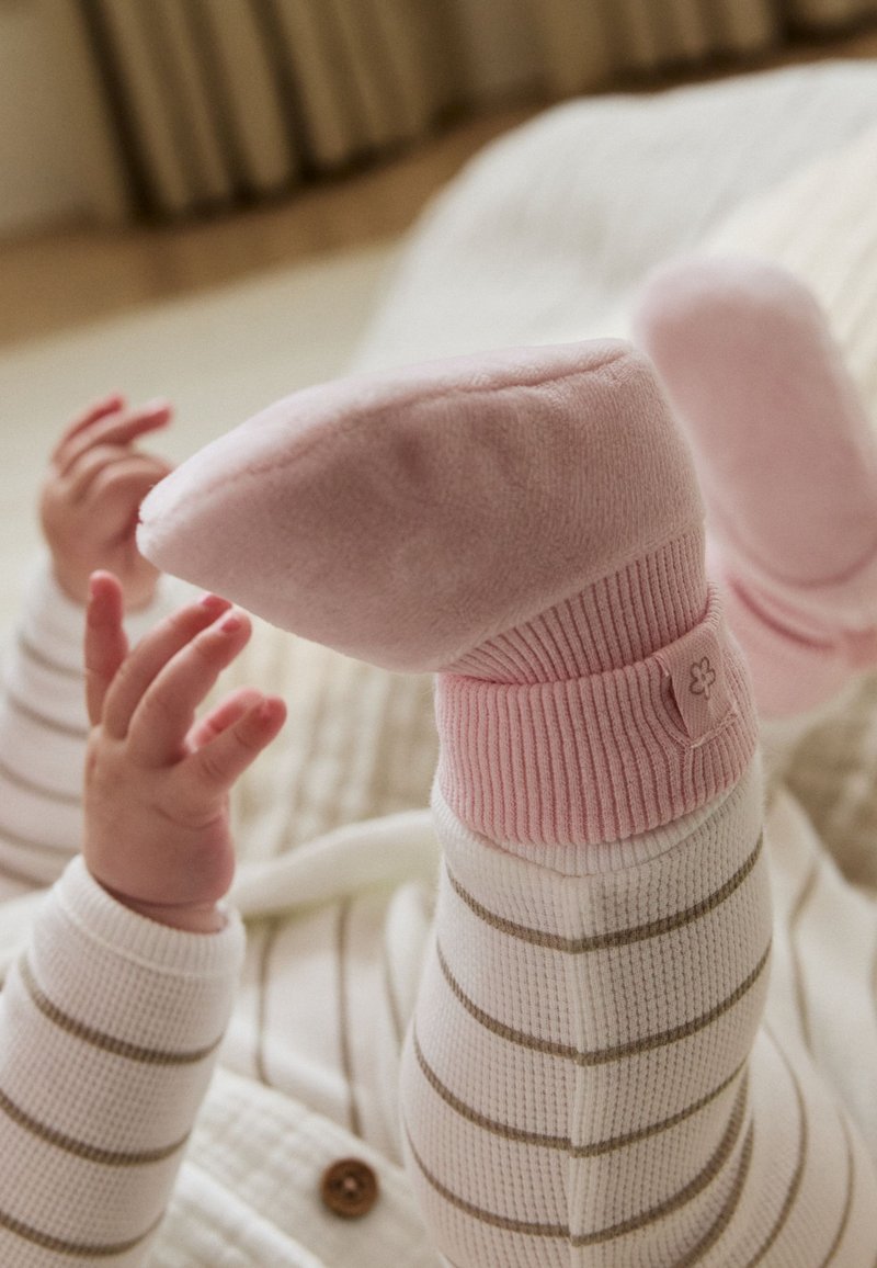 Bébé portant des chaussettes roses et un pantalon blanc rayé, allongé sur une surface douce avec les mains qui atteignent les pieds.
