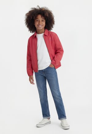 Barn med krøllet hår, smilende, iført rød jakke, hvid t-shirt, blå jeans og hvide sneakers, stående med den ene hånd i lommen.