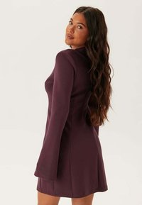 Mini abito aderente a maniche lunghe in tessuto viola scuro. Presenta un collo alto e maniche svasate. Texture liscia con una silhouette semplice.