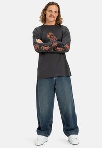 Černé tričko s dlouhými rukávy a oranžovými grafikami na rukávech, v kombinaci s volnými modrými denimovými džíny a bílými teniskami. Neformální streetwear styl.