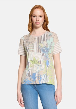 Junge Frau trägt ein beige-, blau- und grün gemustertes T-Shirt mit floralem Muster, gestreiften Ärmeln und blauen Jeans, stehend vor einem einfarbigen Hintergrund.