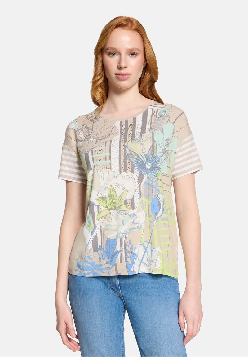 Jeune femme portant un T-shirt à motifs floraux beige, bleu et vert avec des manches rayées, et un jean bleu, debout devant un fond uni.